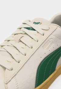 Κοντινό πλάνο μπεζ αθλητικού παπουτσιού Puma Suede με πράσινη πλευρική λωρίδα, άσπρα κορδόνια και καουτσούκ σόλα.