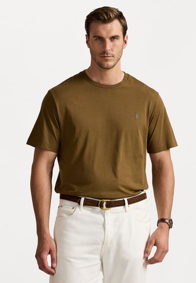 Camiseta de algodón marrón con cuello redondo y mangas cortas. Presenta un pequeño logo bordado en el pecho. Combinada con jeans blancos y un cinturón marrón.
