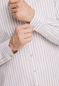 Camisa de algodón a rayas en beige y blanco con cuello abotonado. Destacados botones dorados en los puños y en la tapeta delantera.