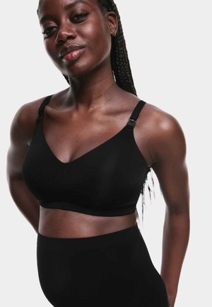 Femme confiante aux cheveux tressés portant un soutien-gorge sans fil noir doux et un bas taille haute noir sur un fond uni.