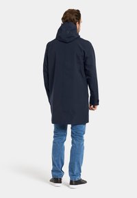 Marineblauer Regenmantel mit Kapuze, gerade geschnitten, mit langen Ärmeln und glatter Oberfläche. Kombiniert mit blauen Jeans und schwarzen Sneakers.