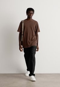Man walking forward wearing brown oversized t-shirt, black pants, white sneakers, and carrying a beige shoulder bag against a plain white wall.