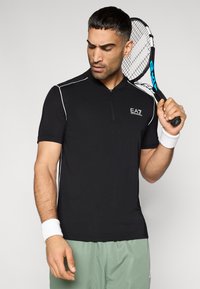 Μαύρο αθλητικό πουκάμισο με κοντό μανίκι και φερμουάρ στο ένα τέταρτο, λευκά στοιχεία και λογότυπο EA7. Το μοντέλο κρατάει ρακέτα τένις και φοράει λευκό βραχιολάκι στον καρπό.