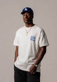 Balta kokvilnas T-kreklu ar zilu izšūtu tekstu "Believe Original Clothing" kreisajā krūtī. Valkāts ar melniem šortiem un kārtām ķēdēm.