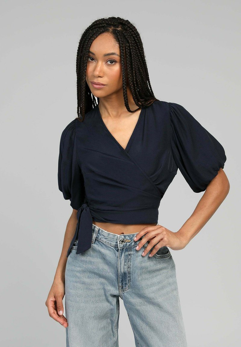 Femme avec de longs cheveux tressés portant un top court enveloppant bleu marine et un jean taille haute bleu clair, devant un fond gris uni.