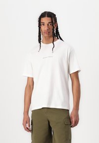 Hvid bomulds-t-shirt med korte ærmer, prydet med teksten "VELKOMMEN TIL PARADIS" trykt i midten. Enkel design, afslappet pasform.