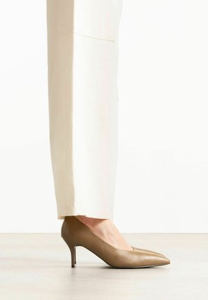 Frau trägt beige spitze Leder-Kitten-Heels kombiniert mit off-white geraden Hosen, steht auf einem hellen Boden.