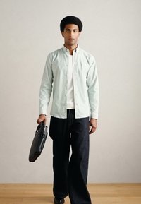 Ανοιχτό μπλε πουκάμισο με κουμπιά, λευκό εσώρουχο, σκούρο τζιν παντελόνι. Το μοντέλο κρατά μια μαύρη τσάντα, στέκεται σε ξύλινο πάτωμα μπροστά σε ένα απλό τοίχο.