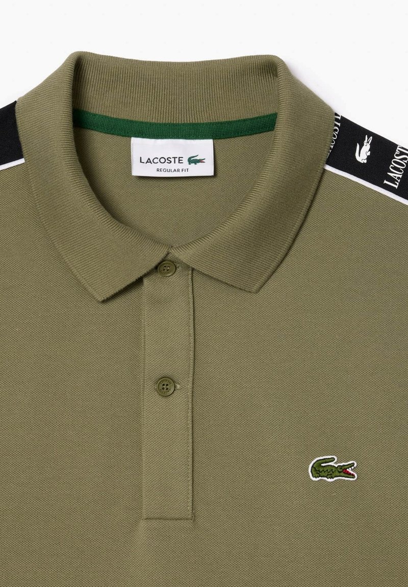 Polo shirt vert olive en tissu texturé, avec deux boutons, un col côtelé et des rayures d'accent noir et blanc sur les épaules. Logo Lacoste présent.