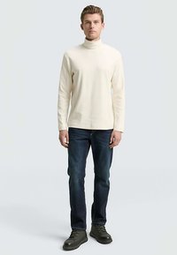 Cream turtleneck shirt, soft texture, long sleeves, paired with dark jeans and rugged black boots. Model stands against a light background.