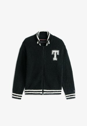 Garçon portant une casquette rouge, une veste noire zippée avec la lettre T, une chemise blanche, un pantalon cargo beige et des baskets beiges, debout avec une main dans la poche et l'autre ajustant sa casquette.