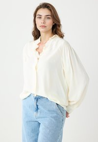 Kremowa bluzka z falbaniastym kołnierzykiem, zapięciem na guziki i bufiastymi rękawami. Noszona z jasnoniebieskimi dżinsami z wysokim stanem. Gładka tekstura.