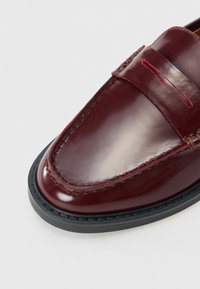 Mocassin en cuir bordeaux avec semelle noire, couture mocassin et une bride avec une découpe décorative rouge.