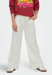 Bijele hlače širokih nogavica s teksturiranim materijalom, s pojasom na vezanje i bočnim džepovima, u kombinaciji s crvenim majicom s grafikom i tenisicama.