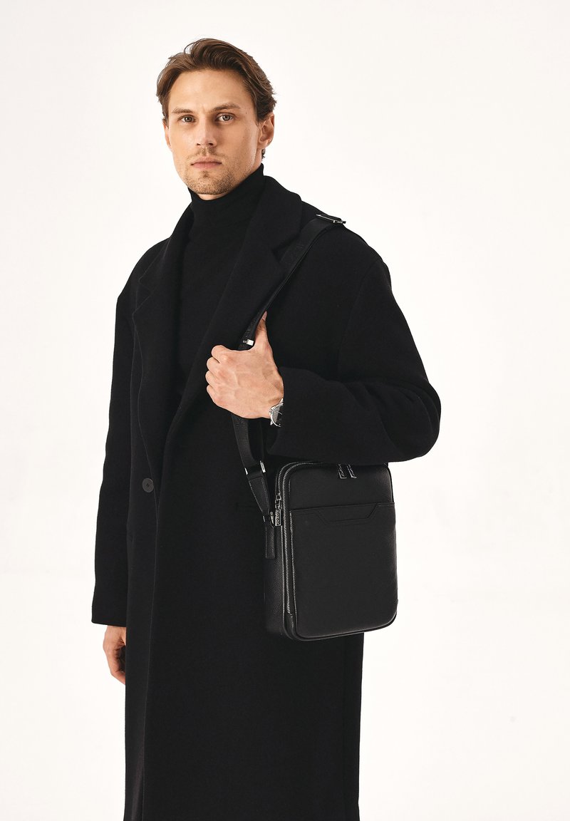 Borsa a tracolla nera in pelle con design strutturato, chiusura con cerniera e tracolla regolabile, abbinata a un lungo cappotto nero e un dolcevita.