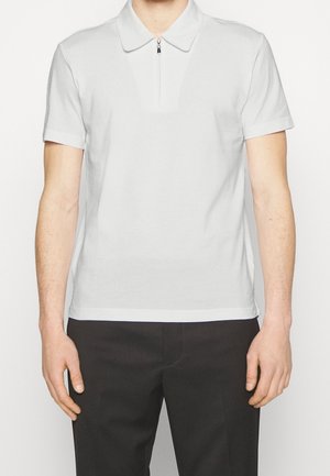 Mężczyzna ubrany w biały krótki rękaw polówkę z przednim zamkiem błyskawicznym, w połączeniu z czarnymi spodniami, na jednolitym tle.