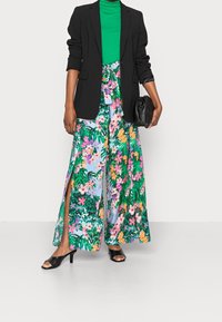 Blazer noir sur un col roulé vert, associé à un pantalon large coloré à fleurs avec fentes et une pochette noire. Sandales à talons noires.