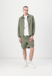 Jachetă verde măsliniu cu fermoar și dungi multicolore, asociată cu pantaloni scurți cargo asortați. Tricou alb și adidași completează ținuta.