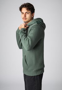 Grüne Kapuzenjacke aus weichem Stoff, mit einer vorderen Tasche, gerippten Bündchen und Saum, sowie dem Schriftzug "BLU" als Akzent auf der Kapuze. Das Modell posiert seitlich.