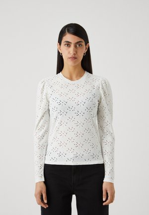 Hvit langermet toppen med intrikate hulldesign, rund hals og subtile pufferm. Kombinert med mørkfargede jeans.