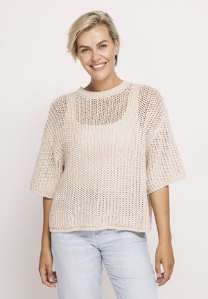 Smiling woman with short blonde hair wearing a loose cream-colored knitted sweater over a tank top and light blue jeans against a plain background.
