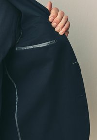 Marineblå blazer i stof med et indre, der har en enkelt lomme med en kontrasterende satintrim og en glat tekstur. Hånd der griber fat om klappen.