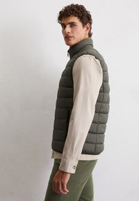 Gepolsterte olivgrüne Weste mit hohem Kragen, Reißverschluss und zwei seitlichen Taschen, getragen über einem hellbeigen Langarmshirt.