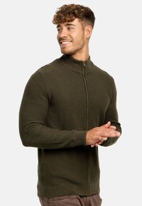 Cardigan de punto verde oscuro con un cuello alto y cremallera frontal. Presenta una tela texturizada y mangas ajustadas, con detalles de ribete en el dobladillo.