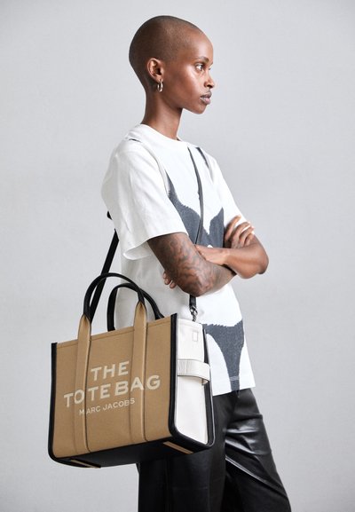 Tan and black tote bag with white accents, featuring a rectangular shape, large text, and sturdy handles. Text: "THE TOTE BAG MARC JACOBS."