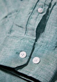 Gros plan d'un poignet de chemise en tissu vert montrant deux boutons blancs et une couture détaillée sur le bord de la manche.