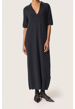 Kobieta ubrana w czarną, krótkorękawową sukienkę sięgającą kostek z głębokim dekoltem w serek oraz czarne szpilki z paskami wokół kostek.