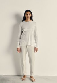 Chemise blanche à manches longues et pantalon de couleur claire, avec une coupe décontractée. Associé à des tongs blanches, sur un fond neutre.