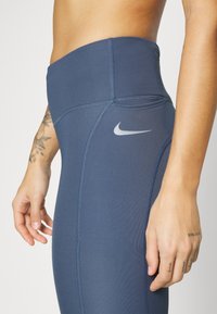 Granatowe legginsy o wysokim stanie z płaskimi szwami i odblaskowym szarym logo Nike z boku. Wykonane z elastycznej tkaniny sportowej.