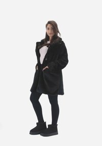 Sort faux fur frakke med sjalskrave, to knapper og sidelommer, parret med sorte leggings og ankelstøvler. Glat tekstur.