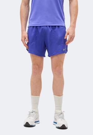 Homme portant un short de sport violet, des chaussettes blanches et des chaussures de course blanches, debout devant un fond clair uni.