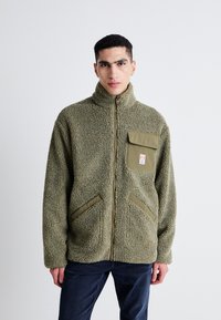 Giacca in pile verde oliva con colletto alto, chiusura con zip, tasca a toppa e due tasche laterali. Tessuto morbido e design comodo.