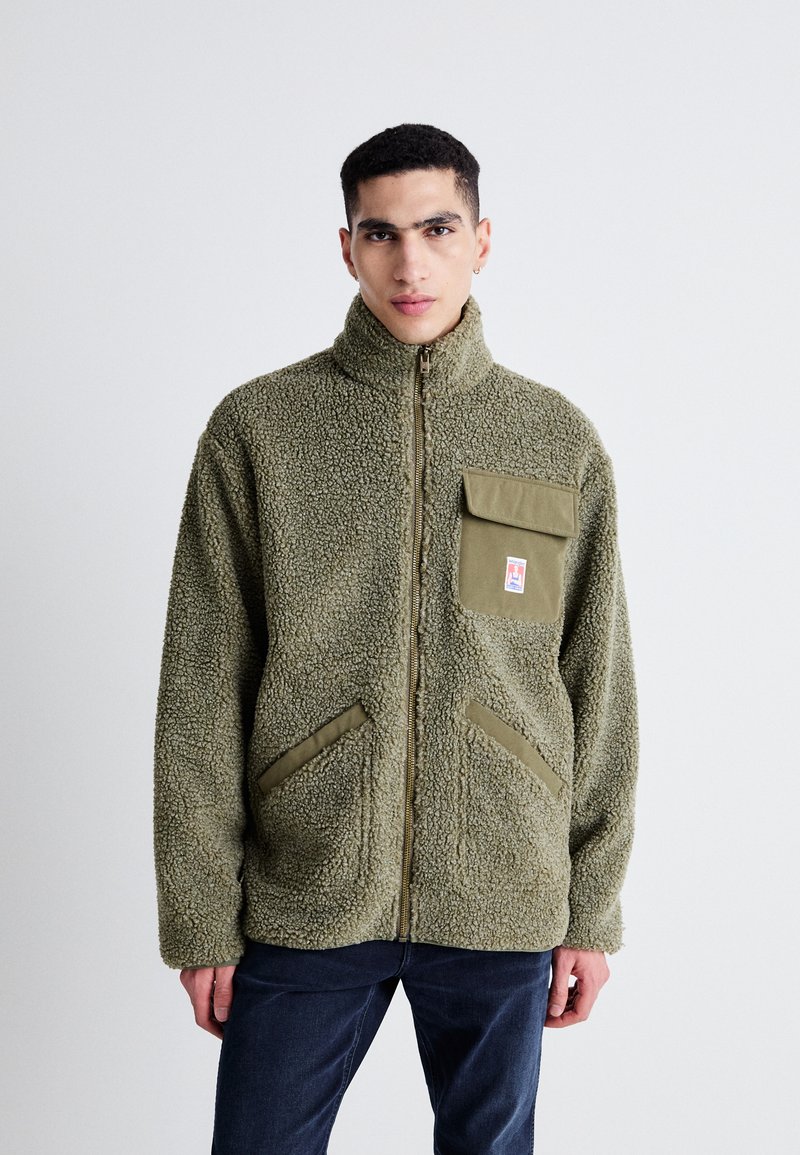 Giacca in pile verde oliva con colletto alto, chiusura con zip, tasca a toppa e due tasche laterali. Tessuto morbido e design comodo.