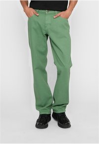 Žalios džinsinės kelnės su tiesiu kirpimu, turinčios priekinės kišenes ir metalinį mygtuko uždarymą. Dėvimos su juodais sportiniais bateliais.