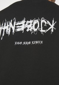 Černá polo triko s krátkým rukávem, které na zádech zdobí asymetrický bílý grafický text s poškozenou texturou a pod ním je umístěna nápis v psacím písmu.