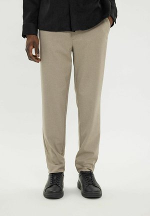 Man die beige op maat gemaakte broek draagt met zijzakken en zwarte leren veterschoenen, staand tegen een effen achtergrond.