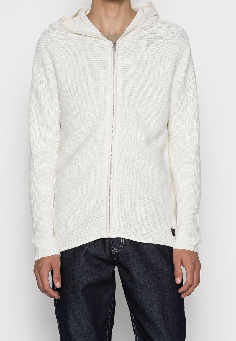 White zip-up hoodie with ribbed texture, featuring a hood and long sleeves, paired with dark denim jeans, shown without additional accessories.