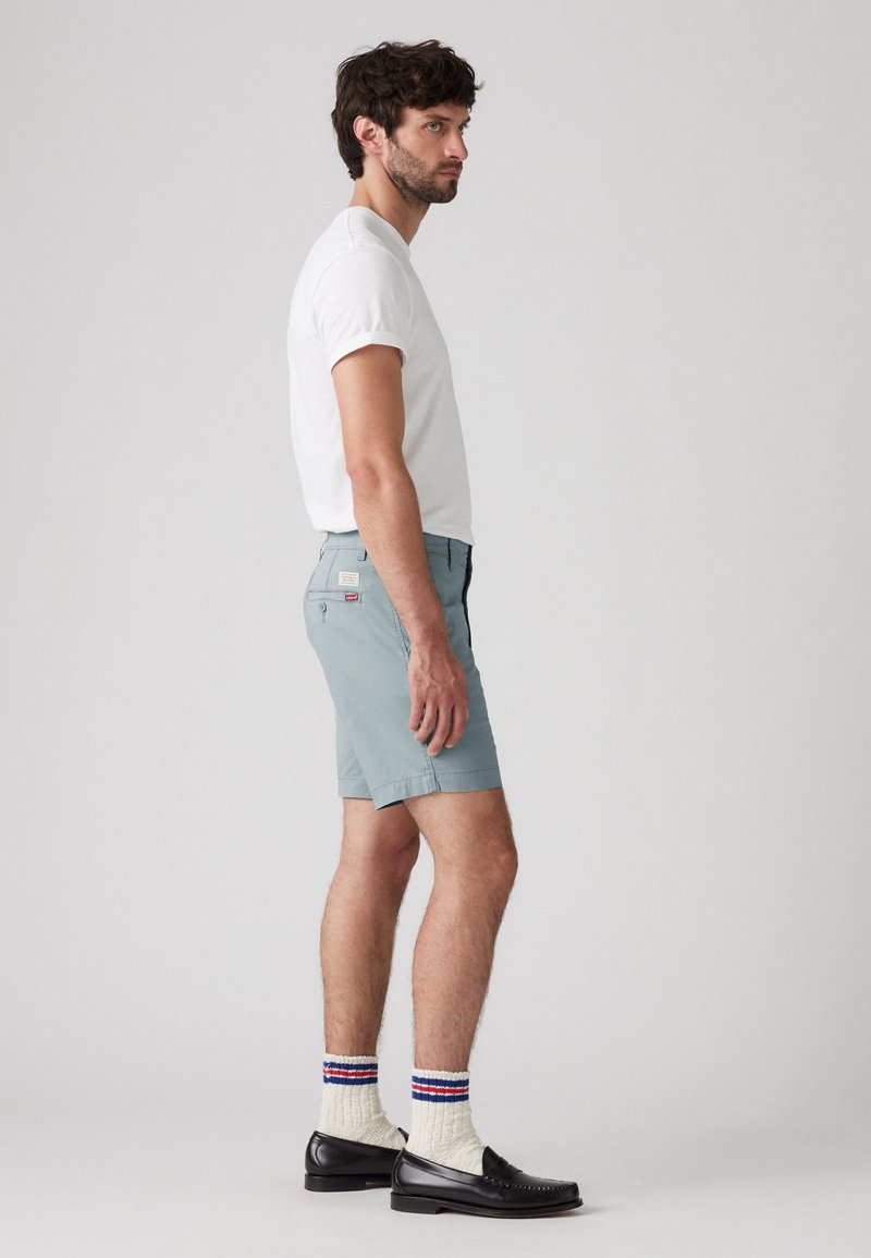 Shorts bleu clair avec une coupe ajustée, portés avec un t-shirt blanc, des mocassins noirs et des chaussettes crème avec des rayures rouges et bleues.