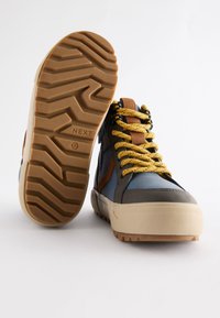 Sneaker alte con tomaia sintetica blu e grigia, dettagli marroni e lacci gialli. Suola in gomma beige spessa con battistrada testurizzato.