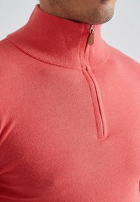 Sweatshirt zippé en corail, avec un col haut et une fermeture éclair d'un quart de longueur. Texture douce avec une finition en tricot lisse.