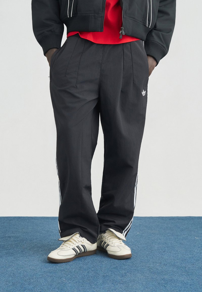 Άτομο που φοράει μαύρο παντελόνι Adidas με λευκές ρίγες και λογότυπο, συνδυασμένο με λευκά παπούτσια Adidas με μαύρες ρίγες, στέκεται σε μπλε χαλί.