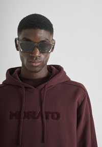 Felpa bordeaux con logo ricamato "MORATO", dotata di cappuccio con cordino. Occhiali da sole chiari e angolari con lenti scure. Texture del tessuto liscia.