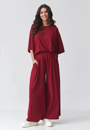 Kobieta z długimi ciemnymi włosami, ubrana w luźny czerwony top i spodnie o szerokich nogawkach, stojąca z rękami w kieszeniach i uśmiechająca się, w białych trampkach.