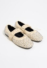 Ballerine beige con una fascia elastica arricciata, adornate con borchie argentate, dotate di punta arrotondata e una fodera interna nera.