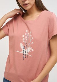 Camiseta de algodón rosa con escote redondo, que presenta un gráfico de pájaros y flores en blanco y azul, y el texto "MUSTANG TRUE DENIM ESTD 1932".