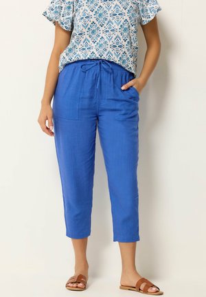 Donna che indossa pantaloni cropped blu brillante con tasche frontali, una maglietta a maniche corte bianca e blu con motivo e sandali marroni a ciabatta.